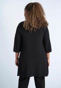 Svart texturerad tunika med trekvartslång ärm, med en löst sittande passform och rak fåll. Bärs över svarta byxor med ett subtilt mönster.