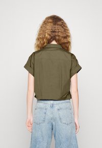 Camicia verde oliva a maniche corte con colletto e maniche arrotolate, abbinata a jeans in denim a gamba larga di colore blu chiaro.