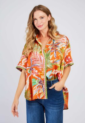 Junge Frau mit welligem blondem Haar, die ein oranges kurzärmeliges Blumenhemd und blaue Jeans trägt, lächelnd mit einer Hand in der Tasche.