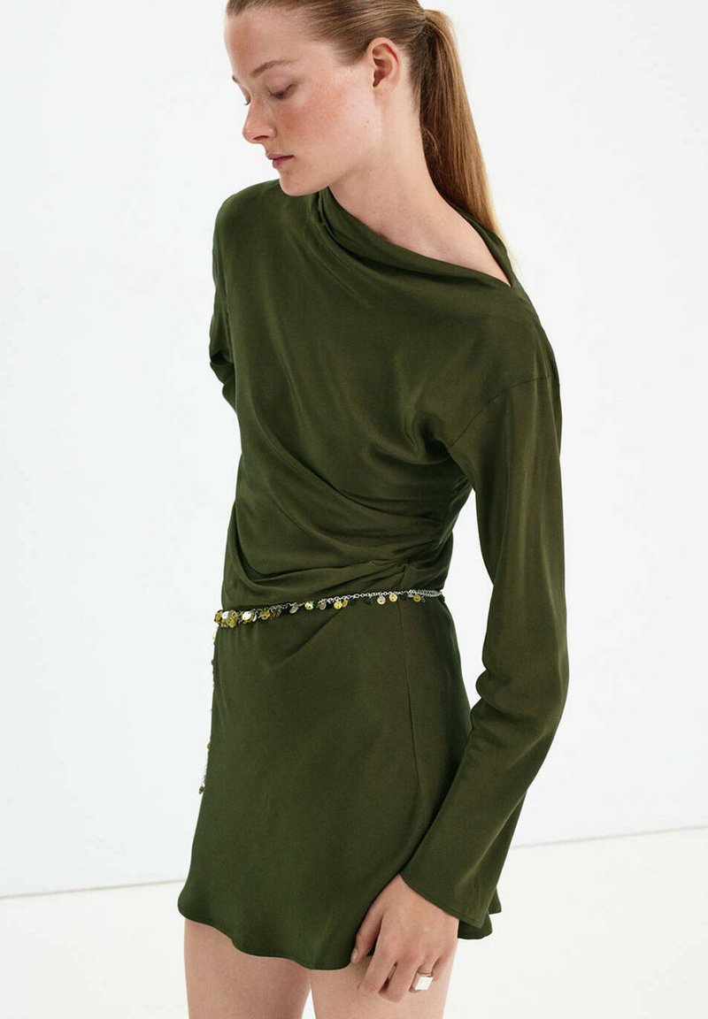 Femme portant une robe à manches longues vert olive avec un décolleté décontracté et une fine ceinture décorative avec des sequins métalliques.