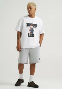 T-shirt bianca oversize con stampa grafica di un personaggio dei cartoni animati e testo, abbinata a pantaloncini di cotone grigi. Il modello indossa scarpe marroni scure.