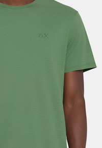 T-shirt verde a maniche corte con un tessuto strutturato. Presenta un piccolo logo blu nella zona sinistra del petto. Collo rotondo e vestibilità normale.