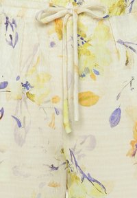 Pantalons en tissu léger à motif floral de couleur crème pâle, avec des accents jaunes et violets. Comprend une taille élastiquée avec cordon.