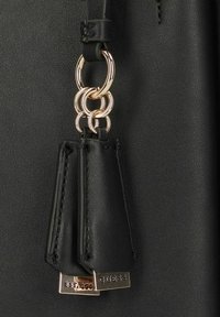 Dettaglio di una borsa a tracolla in pelle nera che mostra una catena in metallo dorato con due nappine in pelle nera, caratterizzata da un'etichetta del marchio con la scritta "GUESS" e "EST. 1981."