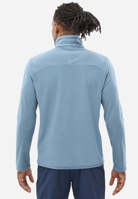 Veste en polaire bleu clair avec un col montant, des manches longues, un tissu texturé et un design simple. Coutures visibles le long de l'ourlet et des poignets.