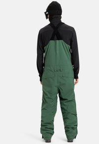 Persoon in groene waterdichte bretelbroek, zwart langemouwen shirt, gebreid muts en zwarte skibril, met gezicht weg van de camera op een witte achtergrond.