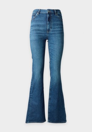 Frau trägt ein schwarz-weiß gestreiftes ärmelloses Oberteil, blaue Jeans mit geradem Bein und schwarze High Heels und steht vor einem schlichten Hintergrund.