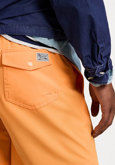 Pantalones cortos naranjas con un bolsillo trasero que presenta una etiqueta, combinados con una chaqueta azul marino. La tela parece suave y tiene un ajuste relajado.