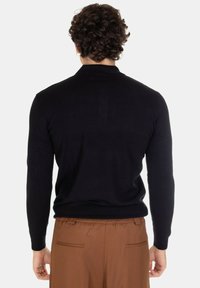 Maglione nero a maglia con maniche lunghe e collo alto, indossato con pantaloni marroni, che mostra una texture liscia e un design aderente.