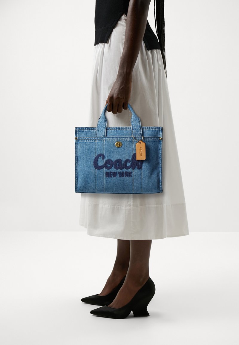 Femme en jupe blanche et haut noir tenant un sac cabas en denim bleu Coach New York avec une étiquette beige et des talons noirs pointus.
