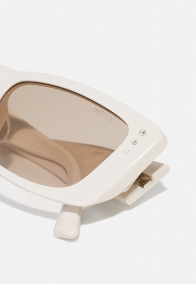 Gafas de sol color crema con un diseño robusto, lentes ovalados y sutiles detalles de piedras de imitación en el marco frontal. Cuenta con una bisagra plegable.