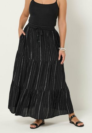 Kobieta ubrana w czarny, bezrękawnik oraz długą czarną spódnicę z srebrnymi pionowymi paskami, w połączeniu z czarnymi sandałami i srebrną bransoletką.