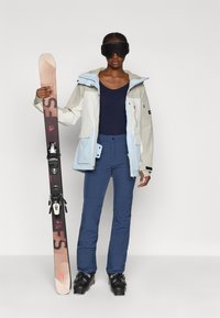 Suksiaisteri, mis sisaldab kahes toonis pehme kanga jakki (helesinine ja valge), tumesinist ülikonda, siniseid isoleeritud pükse ja musti suusasaapaid, kes hoiab suuski.