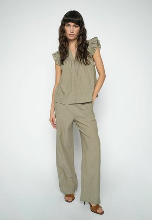 Nő hosszú, hullámos hajjal, olivazöld fodros ujjatlan felsőt, hozzáillő, bő szárú nadrágot és barna nyitott orrú magassarkút viselve, fehér háttér előtt áll.