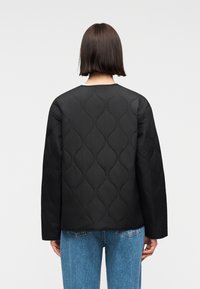 Άτομο με κοντά καστανά μαλλιά, που φοράει μια μαύρη καπιτονέ ζακέτα και μπλε τζιν, με την πλάτη γυρισμένη προς το θεατή, μπροστά από ένα απλό ανοιχτό φόντο.