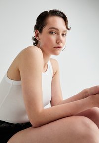 Top alb fără mâneci cu textură cordonată, având un guler rotund. Modelul este așezat, purtând cercei rotunzi din argint, cu ten deschis și într-o poziție casual.