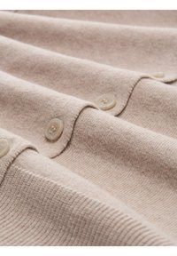 Weiches beige Strickgewebe mit einer Nahaufnahme einer Reihe von durchsichtigen Knöpfen und einem gerippten Saumdetail.