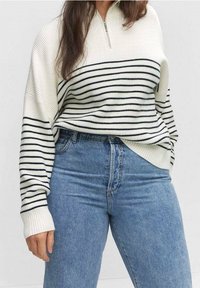 Sweter dziergany z kołnierzem na półzamek, z poziomymi granatowymi paskami na białym tle. Do tego wysokie niebieskie dżinsy z wysokim stanem.