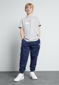 Pilkos spalvos medvilnės marškinėliai su raudonu ir baltu Nike logotipu, sujungti su tamsiai mėlynomis krovinių kelnėmis ir baltais sportiniais batais. Kasdieniškas, patogus stilius.