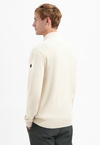 Cream ribbed sweater with a high collar, textured fabric, and fitted cuffs. Features a small logo on the left sleeve.