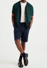 Muž v zeleno-námořnickém vzorovaném kardiganu s krátkým rukávem, bílém tričku, námořnických šortkách, tmavých ponožkách a hnědých kožených botách, stojící s rukama v kapsách.