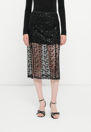 Woman wearing a black long-sleeve top, black sequin-embellished sheer midi skirt, and black ankle-strap sandals with bow and stud details.