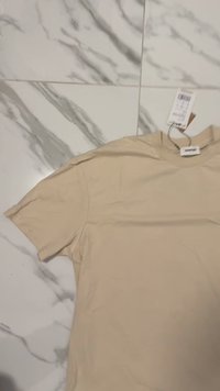 Beige bomulls T-skjorte med korte ermer. Glatt tekstur, rund hals og et merke festet til halslinningen. Lagt flatt på en marmoroverflate.