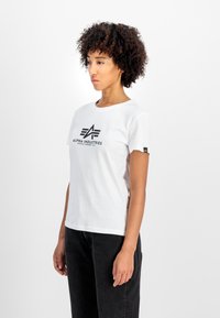 Balta medvilninė marškinėlių viršuje su apvalia iškirpte, su juodu "Alpha Industries" logotipu ir maža geltona prekės ženklo etikete ant rankovės.