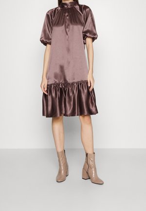 Femme portant une robe ample, mi-longue, brillante, mauve foncé avec des manches bouffantes et des bottines beiges à talons.