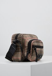 Ruskean ja vaaleanpunaisen ruudullinen kangaskassi, jossa kaksi vetoketjullista taskua, mustat metalliosat ja säädettävä hihna. Pinnan yksityiskohtainen tekstuuri.