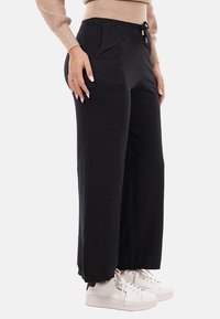 Donna che indossa pantaloni larghi neri con tasche frontali, maglia a maniche lunghe beige e sneakers bianche, in piedi di fronte a uno sfondo bianco.