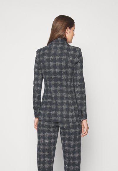 Marineblå ternet skræddersyet jakkesæt med en tætsiddende blazer og bukser. Tekstureret stof med kontrasterende lyse tråde. Har en klassisk krave og knapdetaljer.