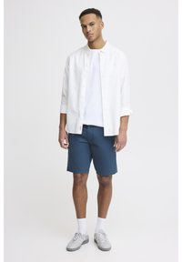 White button-up shirt over a plain white t-shirt, paired with blue shorts. Grey sneakers and white socks complete the outfit.