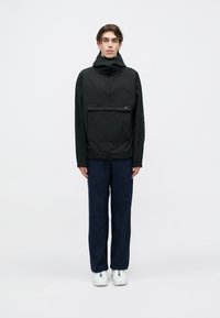 Ungdom som står, iført svart hettegenser med frontlomme, marineblå bukser og hvite sneakers, mot en enkel lys bakgrunn.