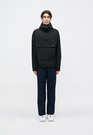 Pessoa jovem de pé, usando uma jaqueta com capuz preta e bolso frontal, calças azul marinho e ténis brancos, contra um fundo liso claro.