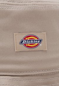 Beige stoffhatt med en flerfarget logo-lapp av "Dickies" med sirkulære aksenter og kontrasterende sømdetaljer.