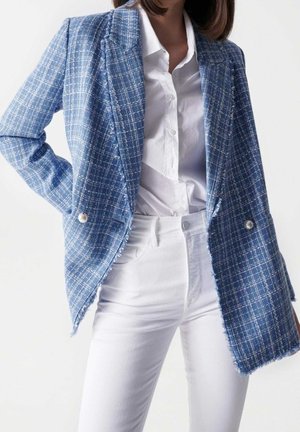 Femme portant une chemise blanche à boutons, un pantalon blanc taille haute et un blazer bleu texturé avec des boutons en perle et des bords effilochés.