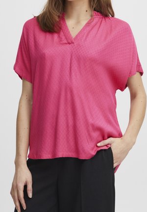 Femme portant un chemisier rose vif à manches courtes avec un motif subtil en forme de losange et un pantalon noir, debout avec une main dans la poche.
