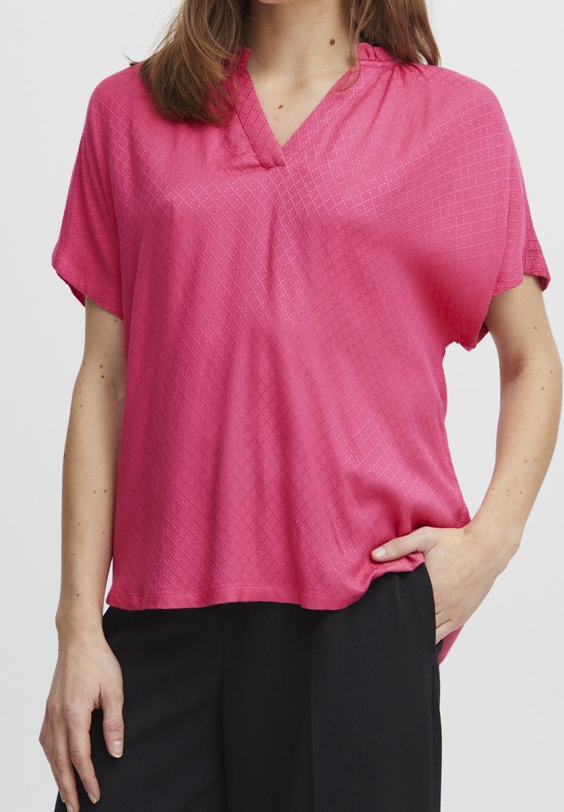 Femme portant un chemisier rose vif à manches courtes avec un motif subtil en forme de losange et un pantalon noir, debout avec une main dans la poche.
