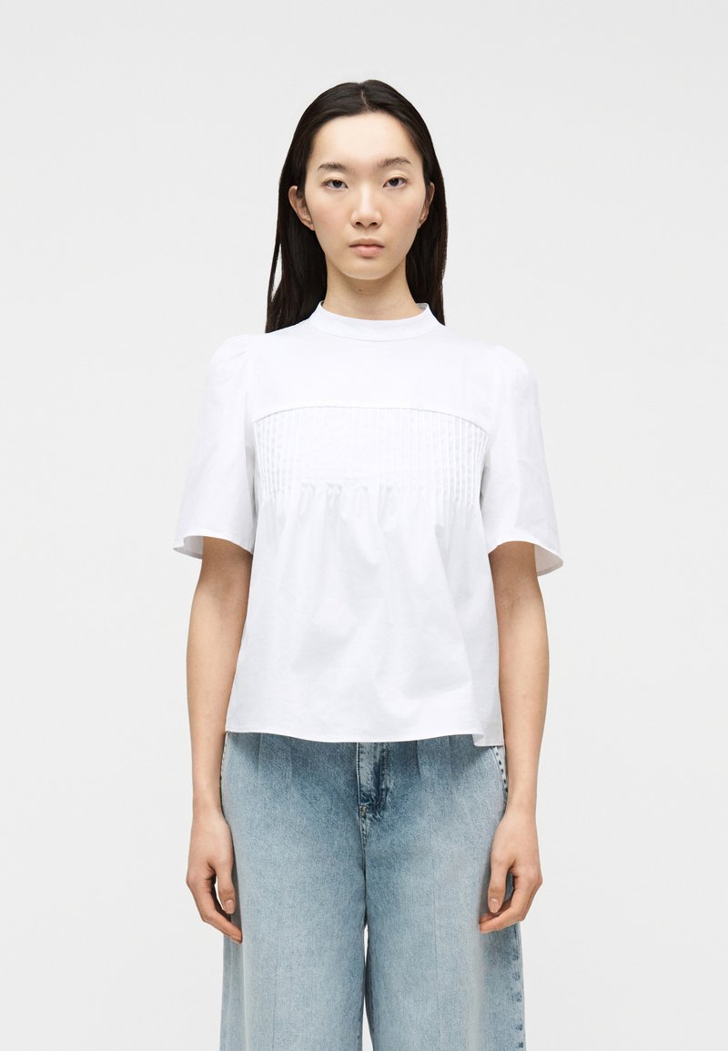 Νέα γυναίκα με μακριά σκούρα μαλλιά φοράει λευκό μπλουζάκι με κοντά μανίκια και λεπτομέρειες με πιέτες, σε συνδυασμό με ανοιχτόχρωμο τζιν ψηλόμεσο, στέκεται σε ουδέτερη στάση.