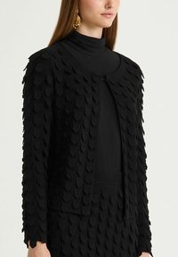 Cardigan noir avec un motif texturé de coquilles superposées. Il présente un col rond et une fermeture à l'avant, porté sur un col roulé noir.