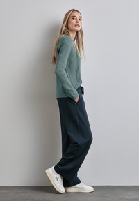 Ljusgrön ribbad stickad tröja, avslappnade mörkblå byxor och vita sneakers. Modellen står med händerna i fickorna mot en neutral vägg.