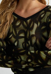 Läpinäkyvä musta neule, jossa vihreä ja kultainen abstrakti kuvio, V-kaulus ja kimaltelevia yksityiskohtia sisältävä teksturoitu kangas.