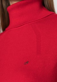 Rode gebreide coltrui met geribbelde structuur en subtiele naaddetails. Heeft een klein zwart logo op de borst.