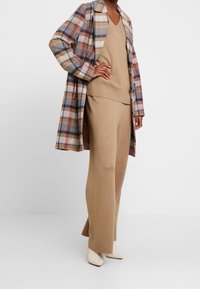 Modèle portant une jupe en tricot beige et un pull à col V associé à un long manteau à carreaux et des bottes blanches à bout pointu, se tenant avec la main sur la taille.