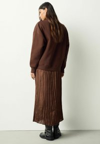 Frau mit langen Haaren, die einen dunkelbraunen übergroßen Pullover, einen plissierten Midirock und schwarze Stiefel mit Schnallen trägt, mit dem Rücken zu einer schlichten Wand stehend.