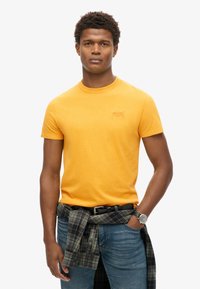 Kortærmet t-shirt med rund hals i klar gul bomuld; har et lille logo på brystet; parret med tætsiddende jeans og en ternet skjorte knyttet omkring taljen.