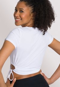 Top corto bianco a coste con arricciature laterali e dettaglio a fiocco in vita. Caratterizzato da un collo arrotondato e una vestibilità slim. Abbinato a pantaloni neri.