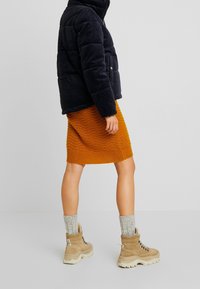 Person som bär en svart vadderad jacka, orange stickad kjol, tjocka grå strumpor och beige vandringskängor, stående mot en enfärgad bakgrund.