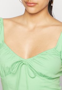 Top verde em algodão com decote em V, mangas franzidas e detalhe de laço na frente. Textura suave com silhueta ajustada.